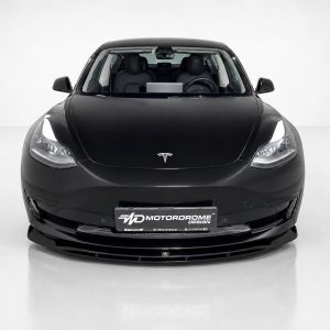 Front view of a black Tesla car in a studio, showing the hood, windshield, and headlights, with a Motordrome Design license plate on the bumper.