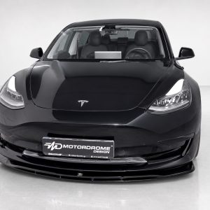 Front view of a glossy black Tesla, headlights on, in a showroom with a 'Motorodrome' license plate.