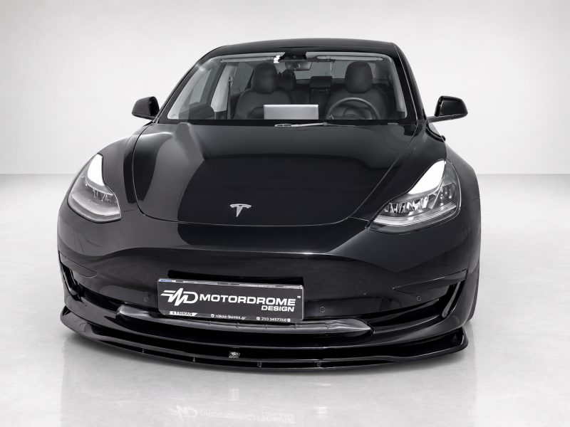 Front view of a glossy black Tesla, headlights on, in a showroom with a 'Motorodrome' license plate.