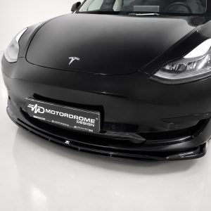 Close-up of a glossy black Tesla front end in a studio, showing headlights and a license plate that reads MOTORDROME DESIGN digitally fainted.