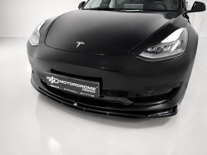 Close-up of a glossy black Tesla front end in a studio, showing headlights and a license plate that reads MOTORDROME DESIGN digitally fainted.