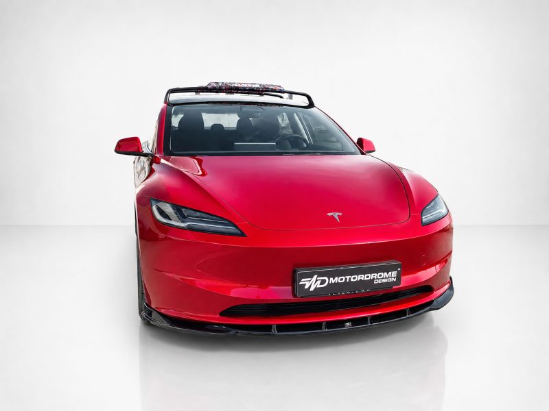 Red Tesla sports car front view with roof rack and visible Tesla emblem, on a white studio backdrop (Motordrome Design plate).