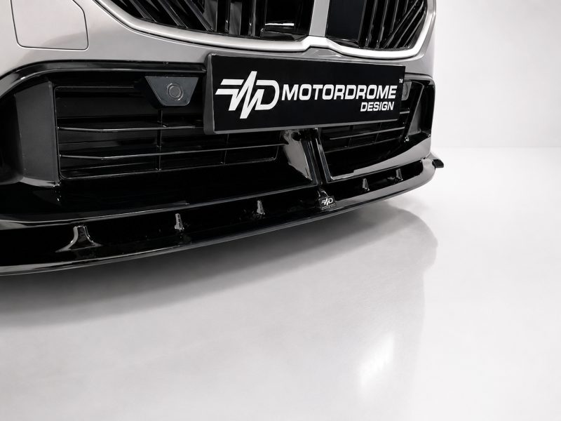 Close-up of a car's black front bumper and grille with a MOTORDROME DESIGN license plate captioned plate in the center.