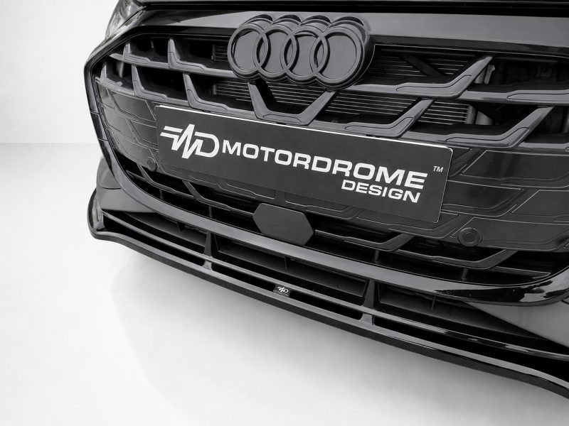 Close-up of a black Audi front grille with the four-ring logo and a Motordrome Design plate in the center, glossy chrome accents evident.