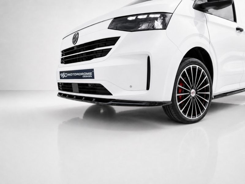 Close-up of a white car's front end showing a black grille, a MotorDrome Design plate, and a multi-spoke wheel with red brake calipers.
