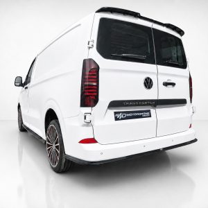 Rear three-quarter view of a white Volkswagen Transporter van in a bright studio, showing red taillights and aftermarket multi-spoke wheels with a MotorDrome badge on the rear.