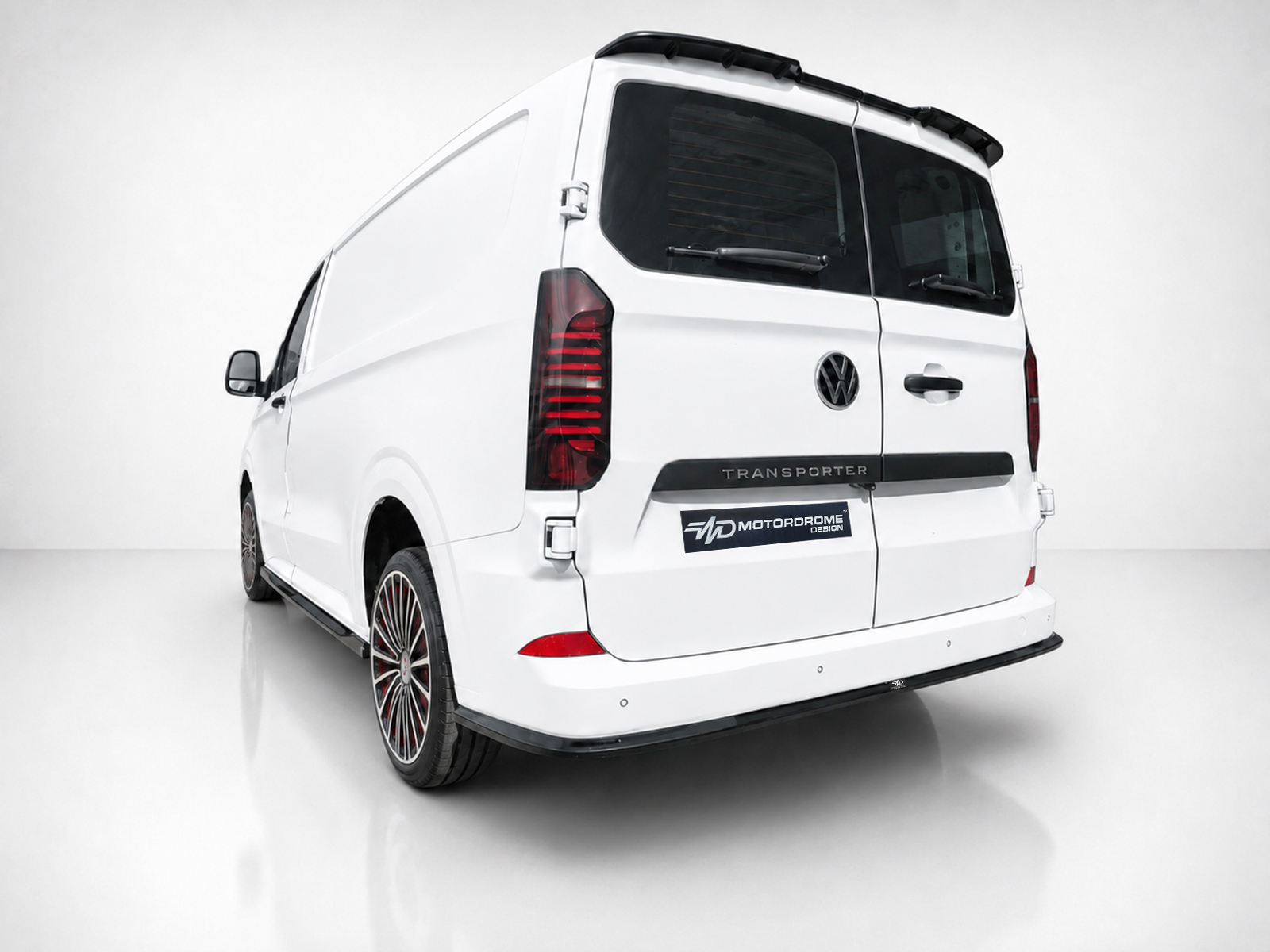 K217-006_1jpg - OPSHOLDERS Rear three-quarter view of a white Volkswagen Transporter van in a bright studio, showing red taillights and aftermarket multi-spoke wheels with a MotorDrome badge on the rear.