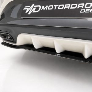 Close-up of a car's front bumper showing a white multi‑fin diffuser and a dark gray bumper with a MOTORDROME DESIGN badge above it, on a light studio backdrop.