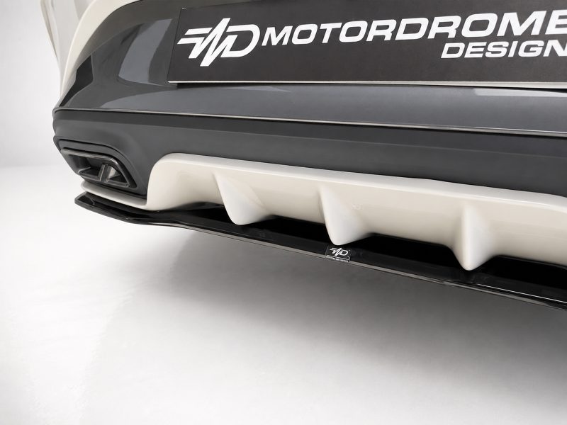 Close-up of a car's front bumper showing a white multi‑fin diffuser and a dark gray bumper with a MOTORDROME DESIGN badge above it, on a light studio backdrop.