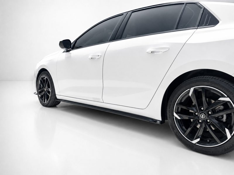 White car photographed from the side in a studio, showing glossy body, black alloy wheels, and tinted windows on a sleek sedan example.