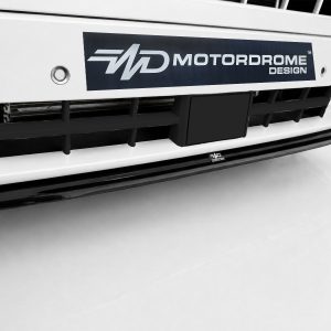 White car front with a dark blue Motordrome Design badge on the bumper and black grille below the tag.