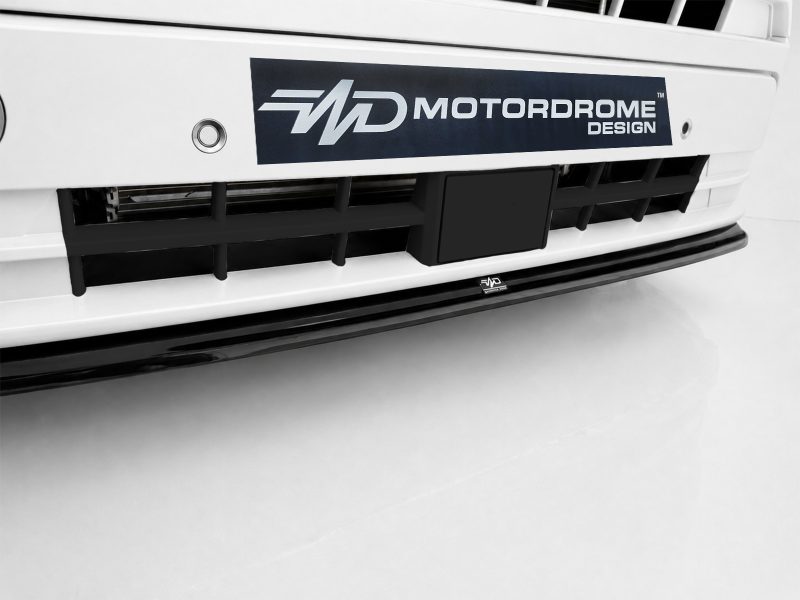 White car front with a dark blue Motordrome Design badge on the bumper and black grille below the tag.
