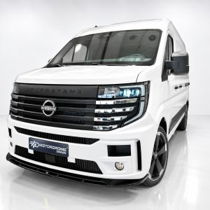 Front three-quarter view of a white Nissan commercial van in a studio, showcasing a bold black grille and blue-tinted headlights with a MotorDrome Design license plate area, on glossy white flooring.