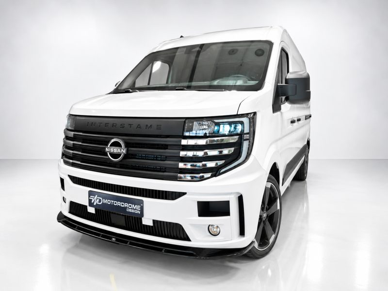 Front three-quarter view of a white Nissan commercial van in a studio, showcasing a bold black grille and blue-tinted headlights with a MotorDrome Design license plate area, on glossy white flooring.