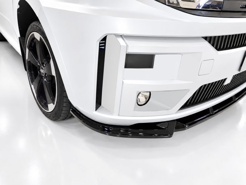 Close-up of a white car's front end, showing the bumper, grille, fog light, and a large black alloy wheel with the Audi emblem visible on the rim