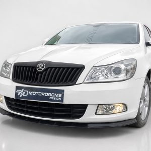 Front-left view of a white sedan showing its black grille, headlight, and a 'Motordrome Design' plate on the bumper.