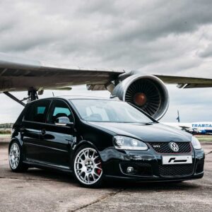 Black VW Golf GTI with aftermarket wheels parked on tarmac beneath a large airplane engine at an airport runway.