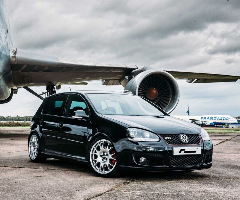 Black VW Golf GTI with aftermarket wheels parked on tarmac beneath a large airplane engine at an airport runway.