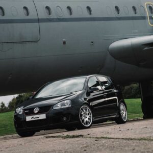 Black VW Golf GTI parked under a large gray airplane fuselage on a grassy field, front angled toward the camera