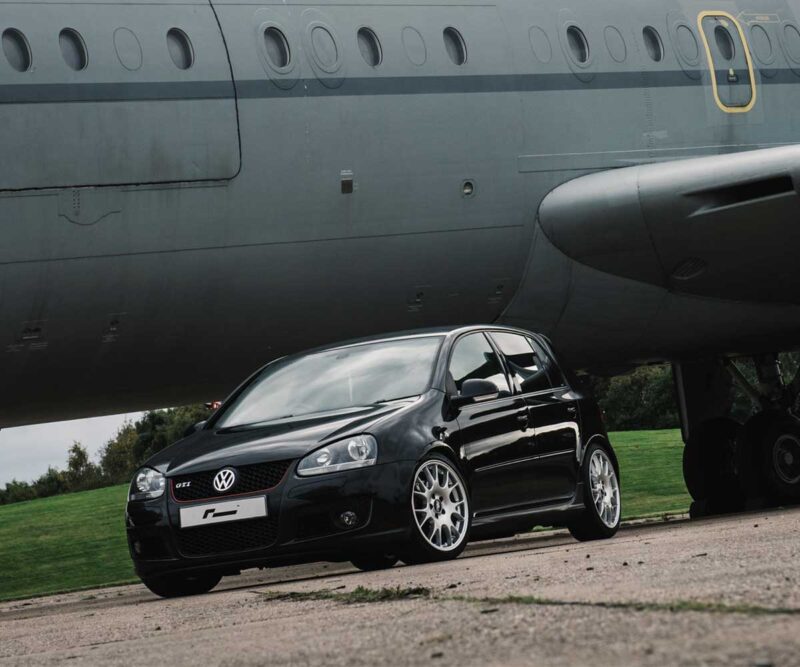Black VW Golf GTI parked under a large gray airplane fuselage on a grassy field, front angled toward the camera