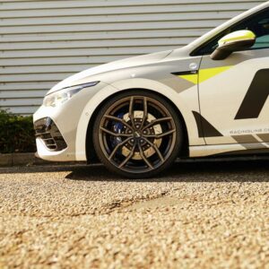 White race car close-up of the front left wheel with blue brake calipers and bold graphic decals on a gravel surface.