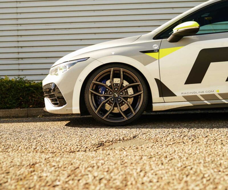 White race car close-up of the front left wheel with blue brake calipers and bold graphic decals on a gravel surface.