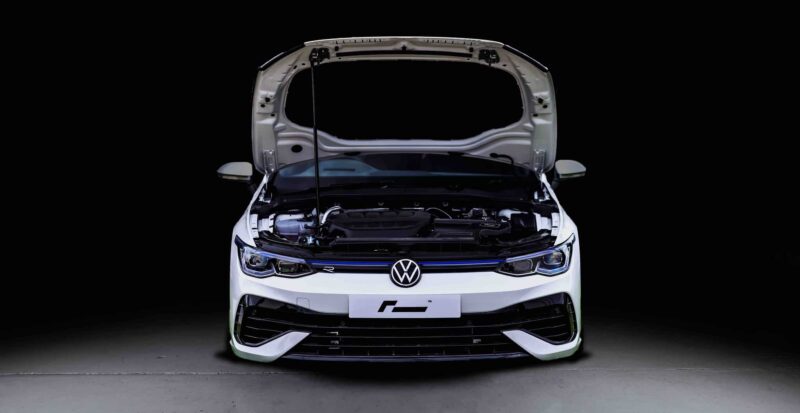 Front view of a white Volkswagen with its hood open, exposing the engine against a black background