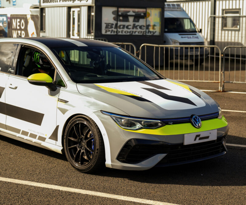 Racingline-Cup-Edition-EVO-Wheels-VWR610010_6-1jpg - OPSHOLDERS White Volkswagen race car with neon yellow accents and bold black graphics parked beside metal barriers at a race track area.