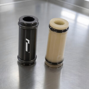 Racingline-DSG-Gearbox-Billet-Oil-Feed-Tubes-DQ500-DQ381-VWR040302_6jpg - OPSHOLDERS Two cylindrical plastic components on a stainless steel surface: one black with a white logo, the other beige with a dark rim.