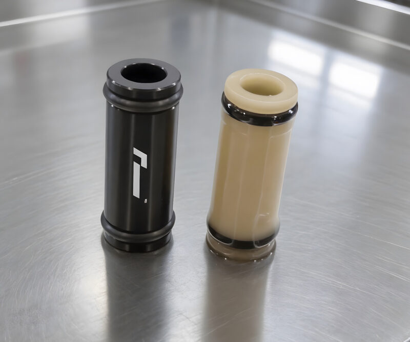 Racingline-DSG-Gearbox-Billet-Oil-Feed-Tubes-DQ500-DQ381-VWR040302_6jpg - OPSHOLDERS Two cylindrical plastic components on a stainless steel surface: one black with a white logo, the other beige with a dark rim.