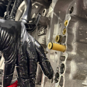 Racingline-DSG-Gearbox-Billet-Oil-Feed-Tubes-DQ500-DQ381-VWR040302_7jpg - OPSHOLDERS Close-up of a mechanic's gloved hand working on a car engine cylinder head, showing yellow fuel/oil passages and surrounding metal gasket area.