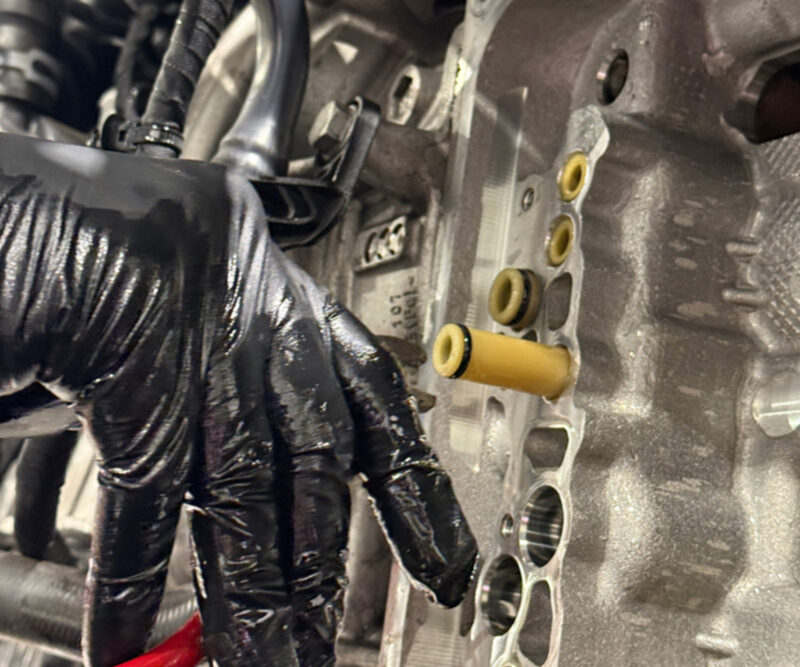 Racingline-DSG-Gearbox-Billet-Oil-Feed-Tubes-DQ500-DQ381-VWR040302_7jpg - OPSHOLDERS Close-up of a mechanic's gloved hand working on a car engine cylinder head, showing yellow fuel/oil passages and surrounding metal gasket area.