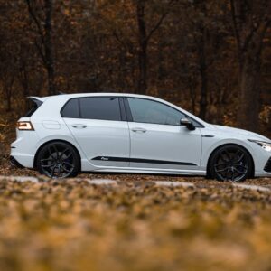 Racingline-Lowering-Springs-Mk8-R-1jpg - OPSHOLDERS White hatchback car parked on a leaf-covered road in a autumn forest, side view showing black wheels and dark-tinted windows.