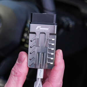 Racingline-PDM-Home-Tuning-VWR031000_5-1jpg - OPSHOLDERS Close-up of a gray metal Power Delivery Module held between fingers, with a white USB-C cable attached against a dark background in a car interior