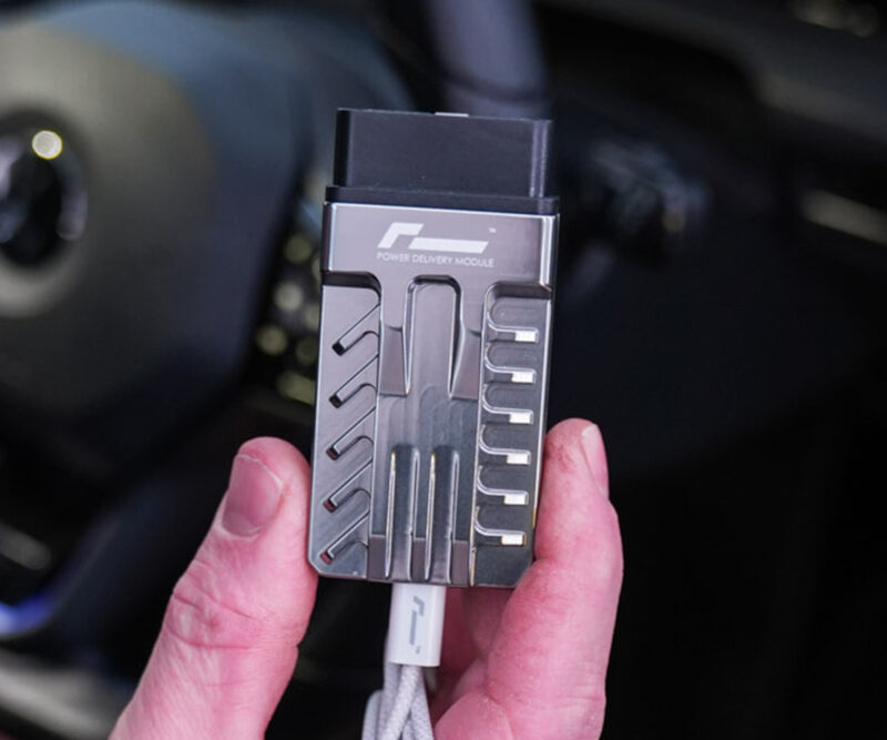 Racingline-PDM-Home-Tuning-VWR031000_5-1jpg - OPSHOLDERS Close-up of a gray metal Power Delivery Module held between fingers, with a white USB-C cable attached against a dark background in a car interior