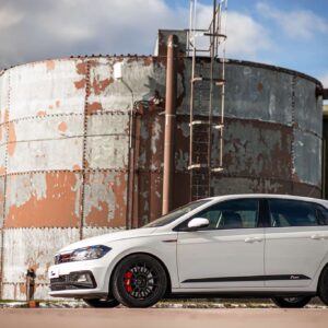 White sedan parked beside a large, rusty cylindrical storage tank with a vertical ladder and corrugated-metal building in the background