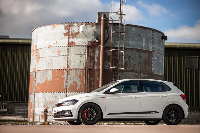 White sedan parked beside a large, rusty cylindrical storage tank with a vertical ladder and corrugated-metal building in the background