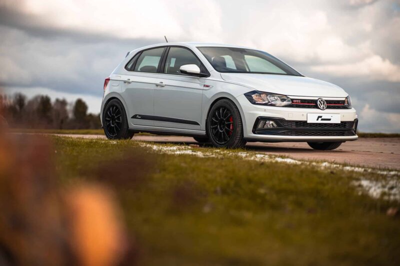 White Volkswagen GTI hatchback parked on a rural road, with a cloudy sky in the background and grass nearby