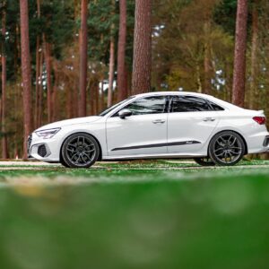 White sedan parked on a paved road with a dense pine forest in the background.