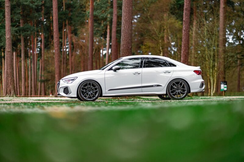White sedan parked on a paved road with a dense pine forest in the background.
