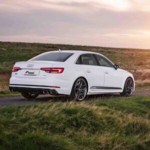 White four‑door Audi sedan on a grassy roadside at sunset, viewed from the rear three‑quarters, with dual exhausts visible.