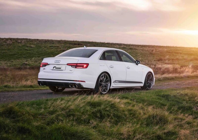 White four‑door Audi sedan on a grassy roadside at sunset, viewed from the rear three‑quarters, with dual exhausts visible.