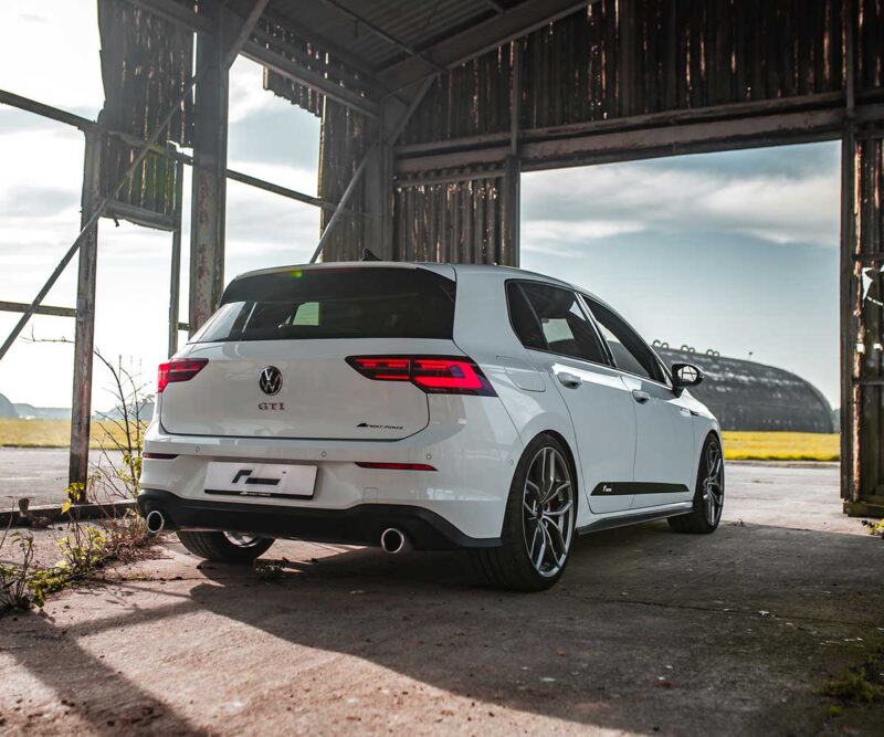 Racingline_MK8_GTIjpg - OPSHOLDERS Rear three-quarter view of a white Volkswagen Golf GTI wagon parked inside a dilapidated shed with rusted metal beams in the background.