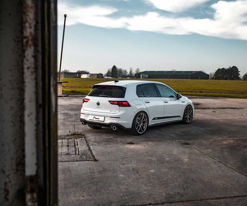 Racingline_MK8_GTI_1jpg - OPSHOLDERS Rear three-quarter view of a white VW Golf GTI Estate parked on a cracked concrete area with open fields and hangars in the distance.