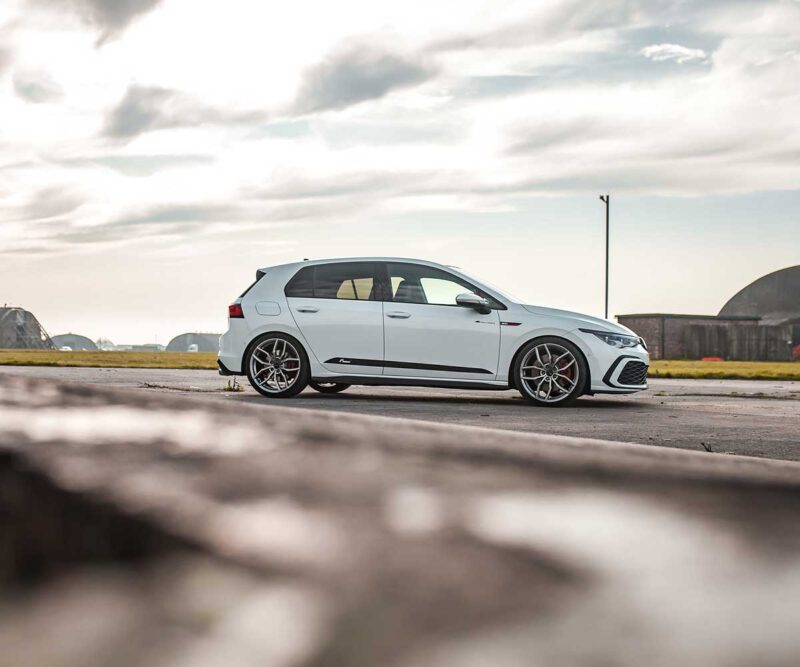 Racingline_MK8_GTI_2jpg - OPSHOLDERS White sporty hatchback parked on an airstrip, with hangars and an open sky in the background.