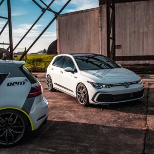 Racingline_MK8_GTI_3jpg - OPSHOLDERS White VW Golf GTI with lowered suspension and large wheels sits on a cracked concrete lot beside a white oem+ hatchback in a rusty industrial yard.