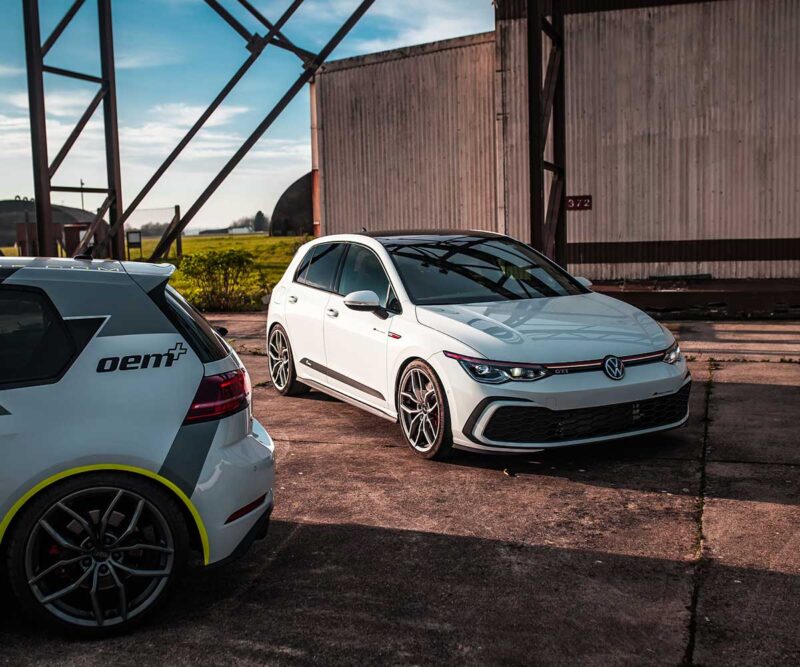 Racingline_MK8_GTI_3jpg - OPSHOLDERS White VW Golf GTI with lowered suspension and large wheels sits on a cracked concrete lot beside a white oem+ hatchback in a rusty industrial yard.