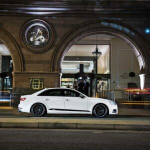 White luxury sedan parked on a city street in front of a hotel entrance at night (The Midland Hotel visible inside).
