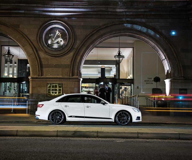 White luxury sedan parked on a city street in front of a hotel entrance at night (The Midland Hotel visible inside).