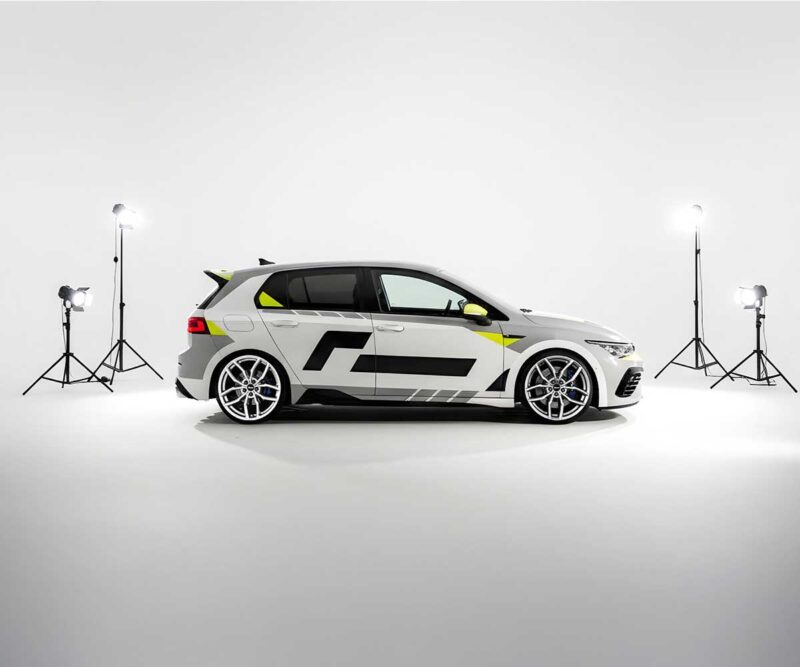 White hatchback car with black racing decals in a white photography studio surrounded by studio lights.